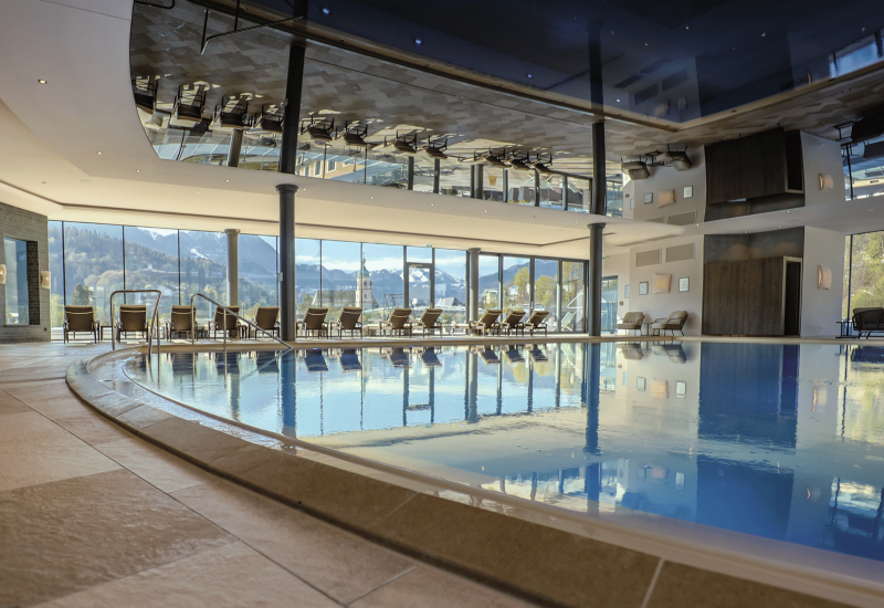 Indoor pool at Hotel EDELWEISS Berchtesgaden with floor-to-ceiling windows, panoramic views of the Alps and comfortable lounge chairs, perfect for a relaxing wellness holiday in Bavaria