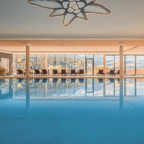 Innenaufnahme eines Schwimmbads mit Blick auf eine Berglandschaft durch große Fenster, Sitze entlang der Fensterfront, Deckenlampe in Form eines Schneeflocke-Designs, ruhige Atmosphäre.