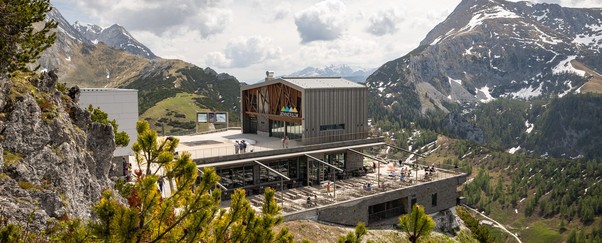 Ein modernes Gebäude auf einer Bergterrasse mit Aussicht auf eine bergige Landschaft mit schneebedeckten Gipfeln, grünen Hügeln und bewölktem Himmel im Hintergrund, umgeben von Kiefern im Vordergrund.