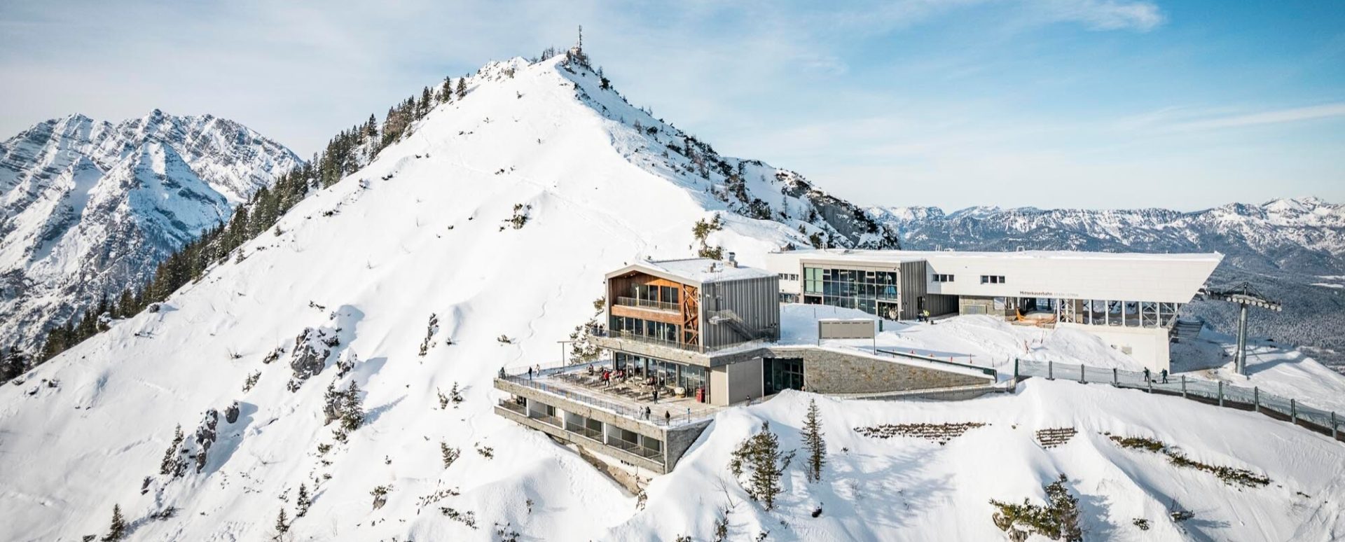Ein modernes Gebäude auf verschneitem Berg, umgeben von hohen, schneebedeckten Gipfeln und einem strahlend blauen Himmel.