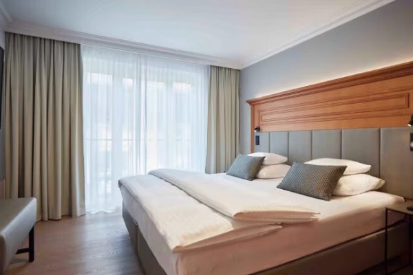 Moderner Hotelzimmer mit großem Bett, grauem Kopfteil, weißen Bettwäsche und zwei grauen Kissen, vor einer großen Fensterfront mit weißen und beige Vorhängen, auf der linken Seite Fernseher und auf der rechten Seite Nachttisch mit Lichtschalter und Telefon.