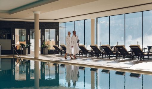 Zwei Personen in Bademänteln spazieren am Pool in einem Wellnessbereich mit großen Fenstern und Poolliegen, Blick auf den Himmel im Hintergrund.