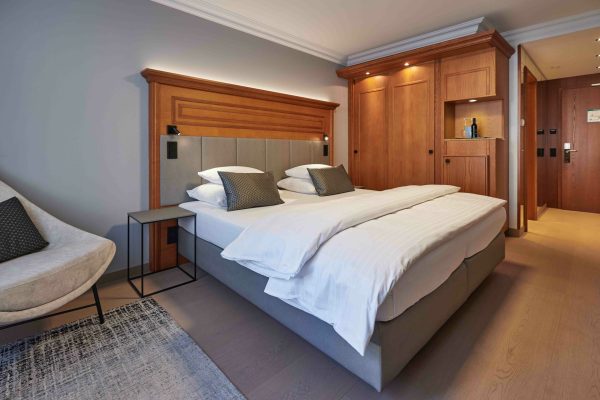 Ein modern gestaltetes Schlafzimmer mit einem großen Bett, das mit weißen Bettwäsche und dunklen Kissen versehen ist, einem beige Lounge-Sessel mit Kissen, holzvertäfelten Wänden und einem eingebauten Holzschrank, sowie Deckenbeleuchtung und Holzfußboden.
