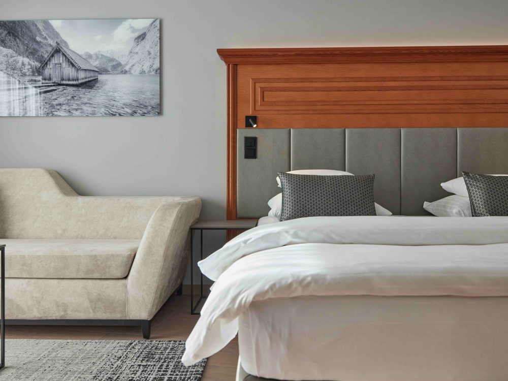 Ein Hotelzimmer mit einem Bett, das an einer aus Holz bestehenden Kopfteil befestigt ist, und einem beigen Sofa auf der linken Seite, an der Wand hängt ein schwarz-weißes Bild eines Hauses an einem See mit Bergen im Hintergrund.