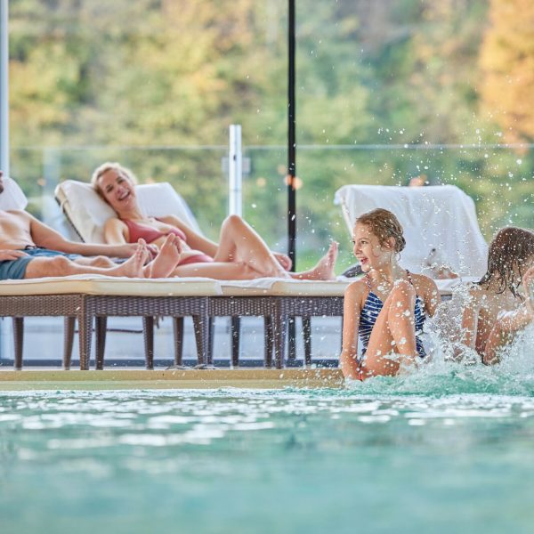 Vier Menschen entspannen sich im Innenbad, zwei Erwachsene auf Liegen im Hintergrund und zwei Kinder im Wasser im Vordergrund, die Spaß haben.
