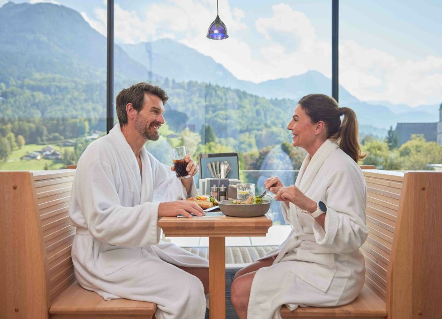 Zwei Menschen in Bademänteln sitzen an einem Tisch in einem Restaurant mit Blick auf eine Berglandschaft, lächeln und essen. Der Mann hält ein Glas Cola in der Hand, die Frau isst aus einer Schüssel.