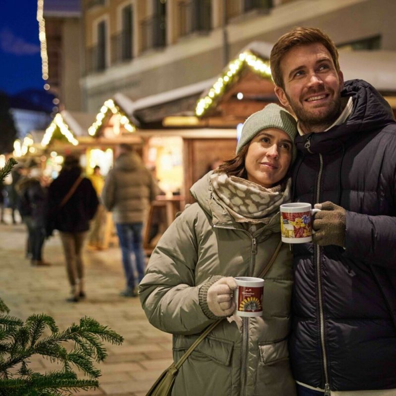 Ein Mann und eine Frau stehen eng beieinander auf einem Weihnachtsmarkt bei Nacht, beide in Winterkleidung, und halten Tassen mit Weihnachtsmotiven, während im Hintergrund festlich beleuchtete Holzhütten und mehrere Menschen zu sehen sind.