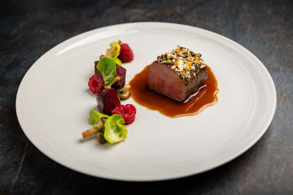 Ein Teller mit einem medaillonförmigen Stück Fleisch, das mit einer Nuss- und Samenkruste bedeckt ist, sitzt in einer braunen Sauce, umgeben von kleinen Beilagen aus Rote Beete, Beeren, Salat und Pilzen auf einem weißen Teller.