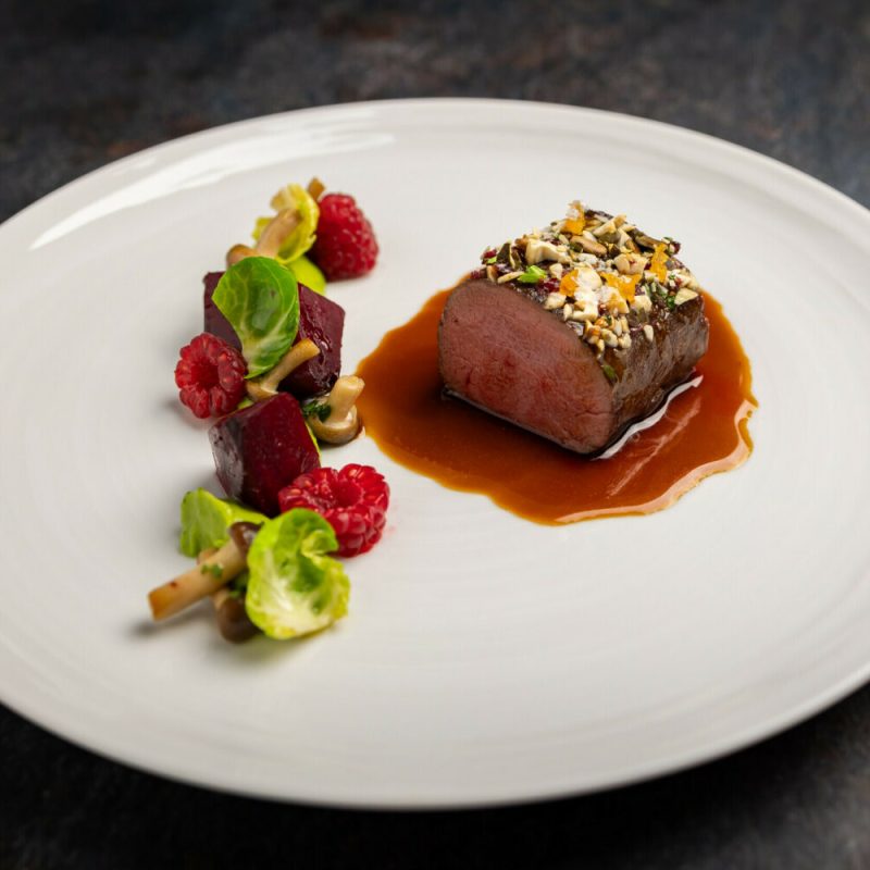 Ein Teller mit einem medaillonförmigen Stück Fleisch, das mit einer Nuss- und Samenkruste bedeckt ist, sitzt in einer braunen Sauce, umgeben von kleinen Beilagen aus Rote Beete, Beeren, Salat und Pilzen auf einem weißen Teller.