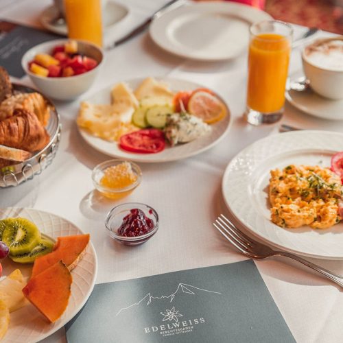 Ein gedeckter Frühstückstisch mit verschiedenen Speisen und Getränken, darunter frisches Obst, Säfte, Joghurt, Marmelade, Käse, gebackene Brötchen, und eine Portion Rührei, auf weißen Tellern und im Hintergrund.