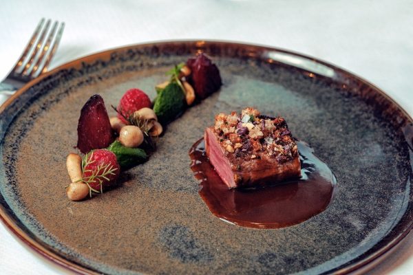 Ein Teller mit einem Stück rosa gebratenem Fleisch, garniert mit einer knusprigen Kruste, liegt in einer dunklen Sauce auf einem dunkelbraunen Keramikteller, daneben befinden sich kleine Kokosnüsse, Himbeeren, eine Tomate, und grünes Gemüse.