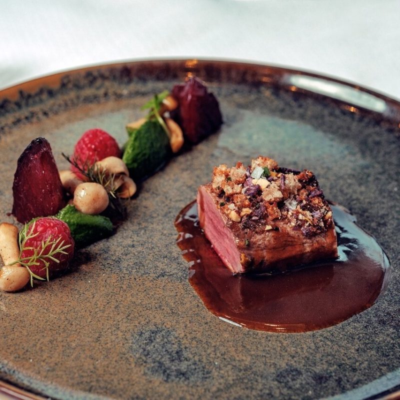 Ein Teller mit einem Stück rosa gebratenem Fleisch, garniert mit einer knusprigen Kruste, liegt in einer dunklen Sauce auf einem dunkelbraunen Keramikteller, daneben befinden sich kleine Kokosnüsse, Himbeeren, eine Tomate, und grünes Gemüse.