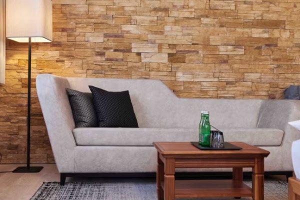 Ein modernes Wohnzimmer mit einem lightgrauen Sofa, zwei schwarze Kissen, einem Holztisch mit Getränkeflaschen und Gläsern, einer Stehlampe mit Lampenschirm und einer Wand aus Steinen in warmen Brauntönen.
