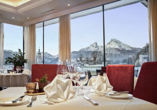 Eleganter Speisesaal mit weiß eingedecktem Tisch, mehrfache Weingläser, Stoffservietten, Geschirr und Besteck, große Fenster mit Blick auf verschneite Berge und Kirchen im Hintergrund.