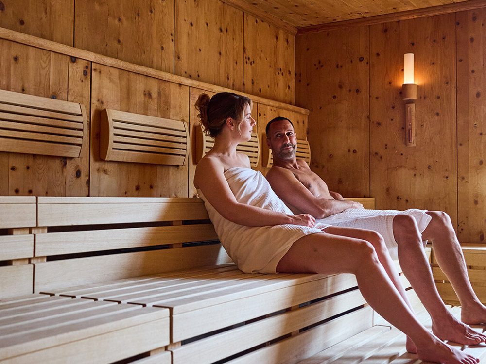 Zwei Menschen, ein Mann und eine Frau, sitzen entspannt nebeneinander in einer Sauna, eingewickelt in Handtücher aus Holz gefertigte Wände und Bänke umgeben sie, mit Sonnenlicht, das durch ein großes Fenster auf die Szene fällt.