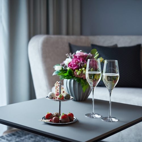 Ein Tisch mit einer Blumenvasen-Arrangement, zwei Gläser Sekt und einer zweistöckigen Dessertplatte mit verschiedenen Süßigkeiten und Beeren in einem modern eingerichteten Raum.