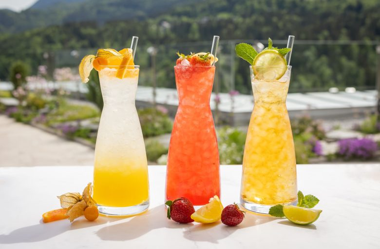 Drei bunte Cocktails in hohen Gläsern mit Früchte Garnituren, darunter eine Zitronenscheibe, Erdbeeren, Limettenscheiben und Minzblätter, auf einer weißen Oberfläche im Freien mit Bergpanorama im Hintergrund.