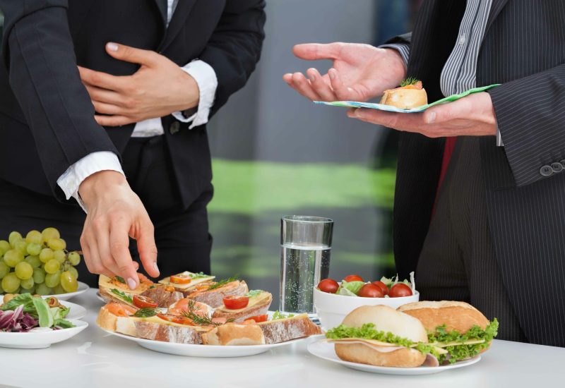 Zwei Personen in formeller Kleidung stehen an einem Tisch mit verschiedenen Sandwiches, Salaten, Trauben, Tomaten und einem Glas Wasser, während eine Person auf der rechten Seite ein Sandwich auf einem Teller serviert.