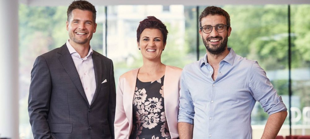 Drei berufstätige Personen, zwei Männer und eine Frau, stehen nebeneinander in einem modernen Büro mit großen Fenstern im Hintergrund, lächeln und schauen in die Kamera.