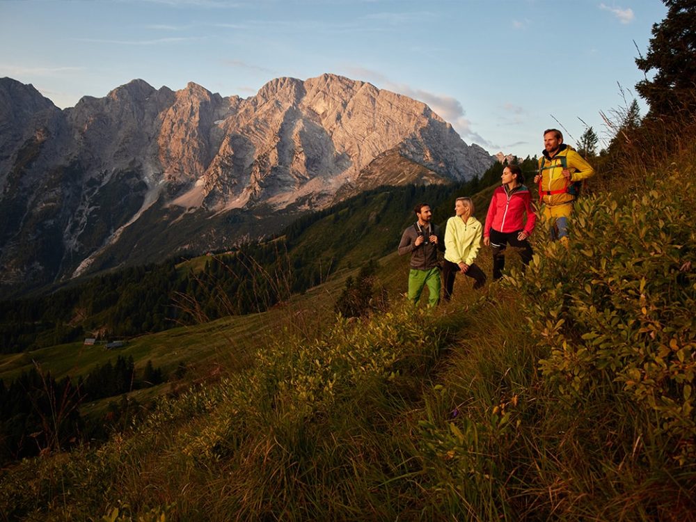 Vier Wanderer auf einem Hügel mit Büschen unterhalb hoher, felsiger Berge bei Sonnenuntergang.
