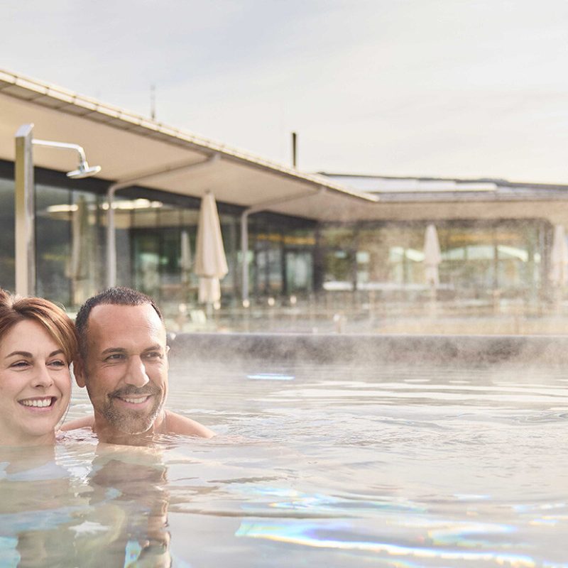 Zwei Personen, ein Mann und eine Frau, entspannen im Außenpool mit Blick auf Berge, während die Sonne aufgeht oder untergeht.