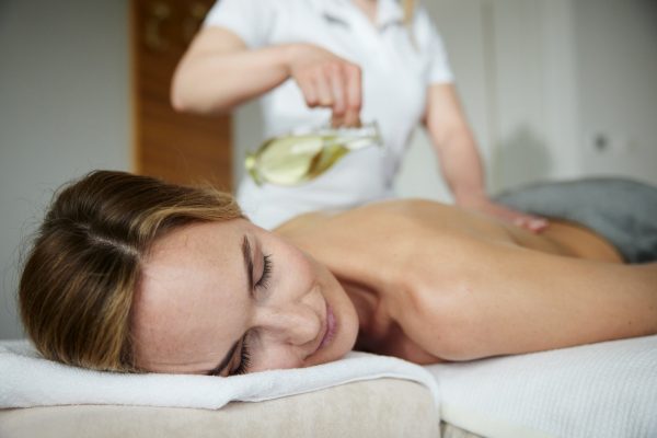 Eine Dame auf dem Bauch liegend wird von einer zweiten Person mit weißem Gewand mit Massageöl massiert