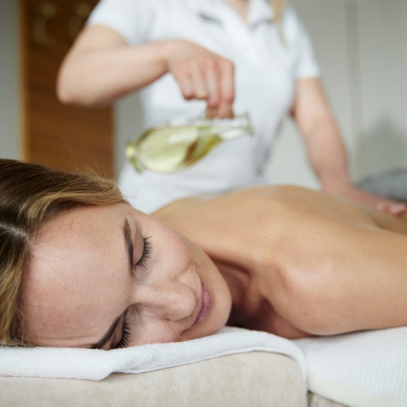 Eine Dame auf dem Bauch liegend wird von einer zweiten Person mit weißem Gewand mit Massageöl massiert
