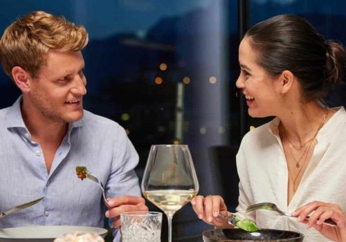 Ein Mann und eine Frau sitzen an einem Tisch bei einem Abendessen, beide lachen und schauen sich freundlich an, mit einem Glas Weißwein zwischen ihnen.