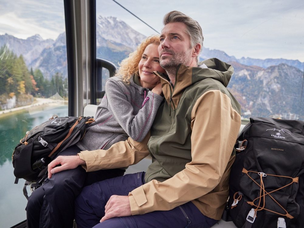 Eine Frau und ein Mann in einer Gondel im herbstlichen Wanderoutfit mit 2 schwarzen Rucksäcken
