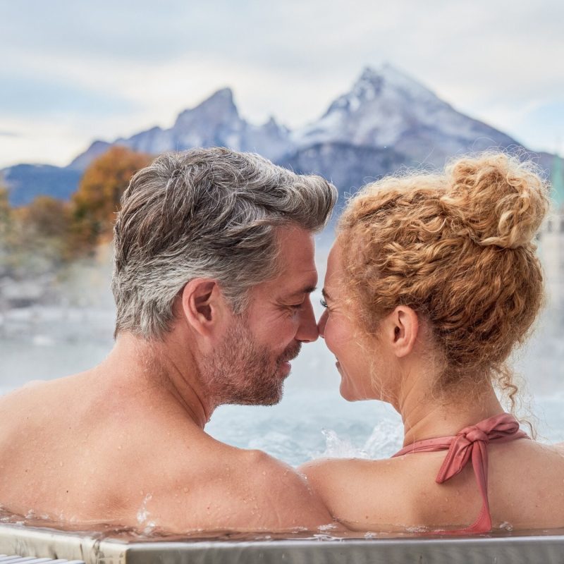 Ein Mann und eine Frau im Whirlpool mit Stirn an Stirn - im Hintergrund ein Berg in bewölkter Stimmung