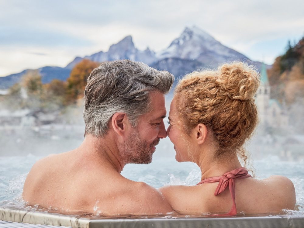Ein Mann und eine Frau im Whirlpool mit Stirn an Stirn - im Hintergrund ein Berg in bewölkter Stimmung