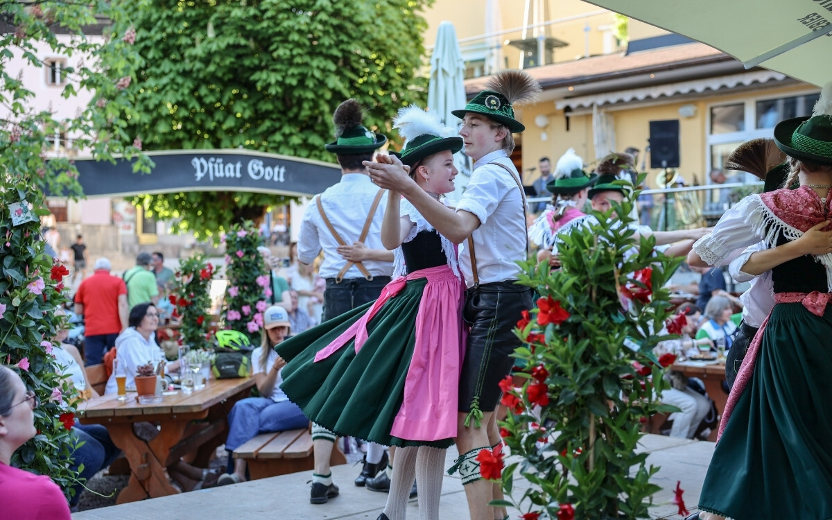 Männer und Frauen in traditionellen bayerischen Trachten tanzen auf einer Bühne bei einem Fest, umgeben von blühenden Blumen und einem Publikum im Biergarten des Gasthof NEUHAUS.