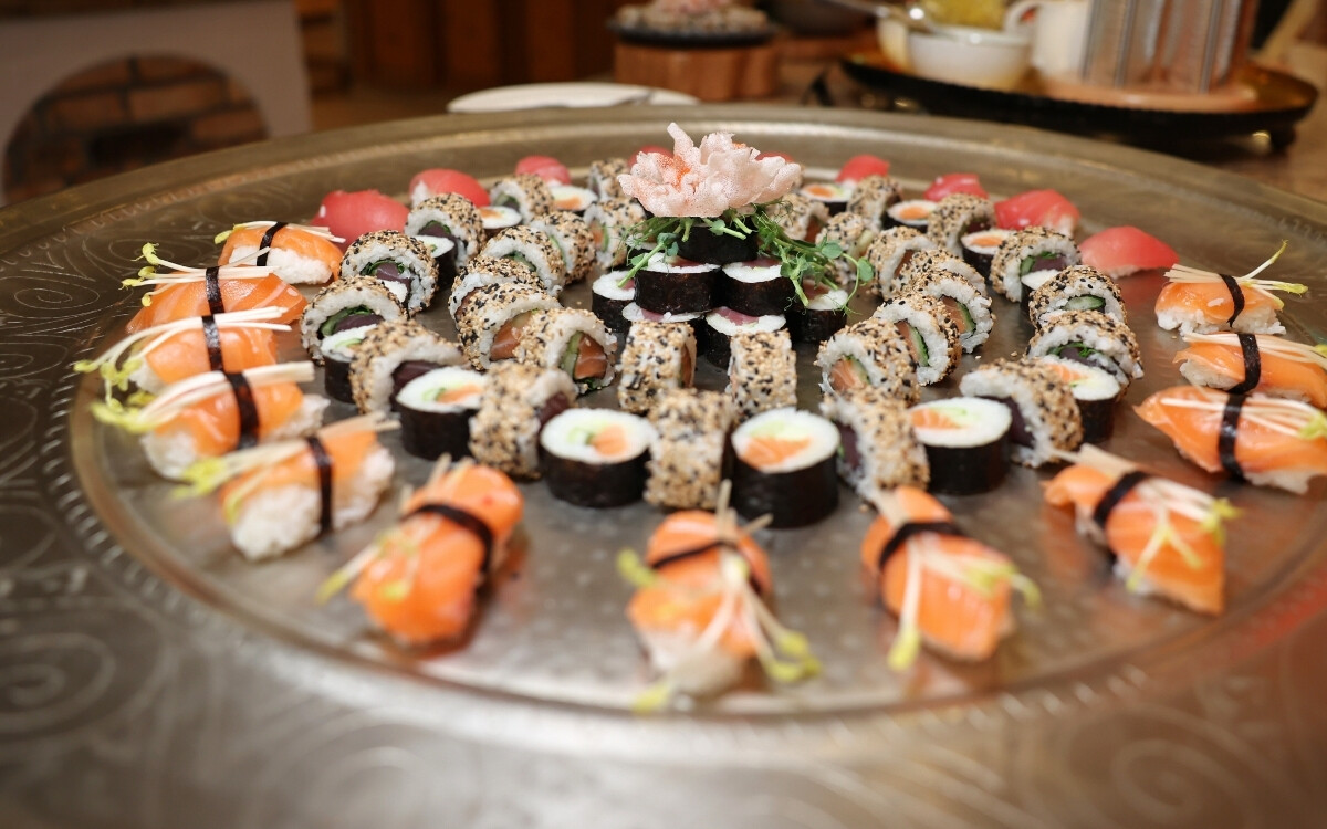 Various sushi rolls and nigiri arranged on a large round silver platter, decorated with a plant in the center, with a well-kept restaurant setting in the background.