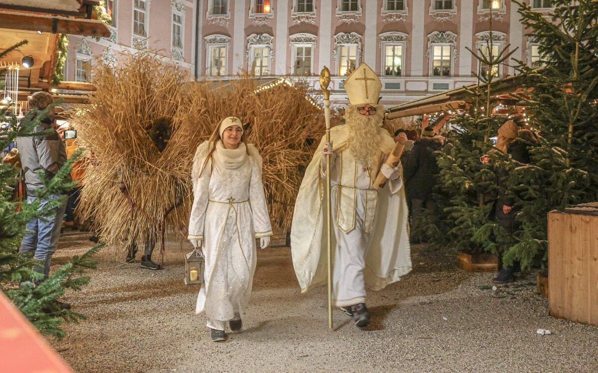 Eine Weihnachtsumzugsgruppe mit einer Person in einem weihnachtlichen Engel-Kostüm, eine andere in einem weißen Märchen-Kostüm, und eine Figur in einem St. Nikolaus-Kostüm, umgeben von Weihnachtsbäumen bei einer festlichen Veranstaltung im Freien.