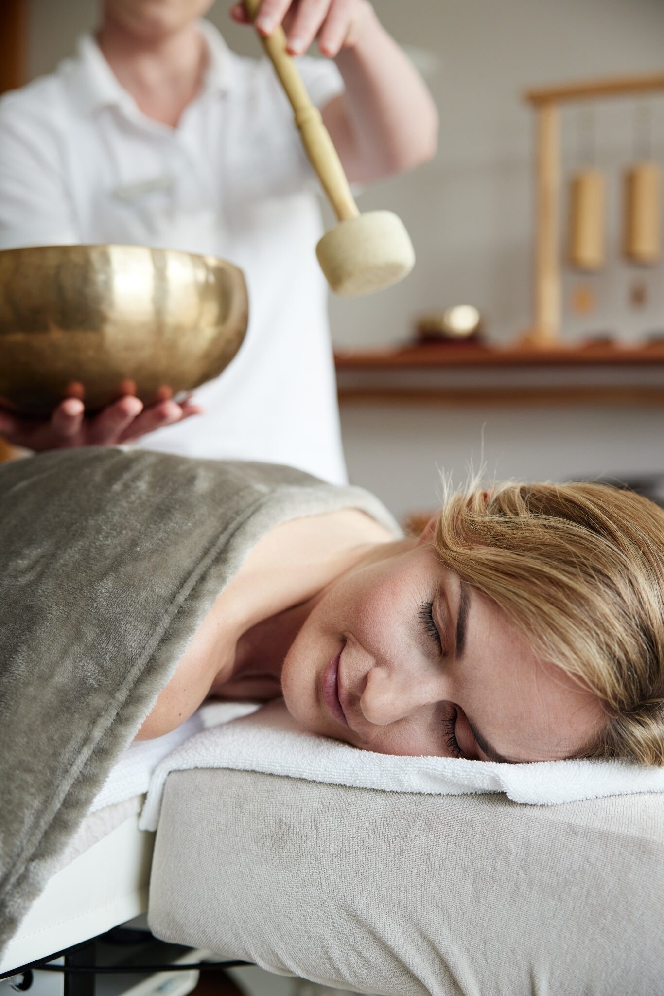 eine Frau liegt entspannt mit geschlossenen Augen auf einer Liege in einer Massagekabine, während eine Masseurin im Hintergrund mit einem Mallet auf eine Klangschale schlägt