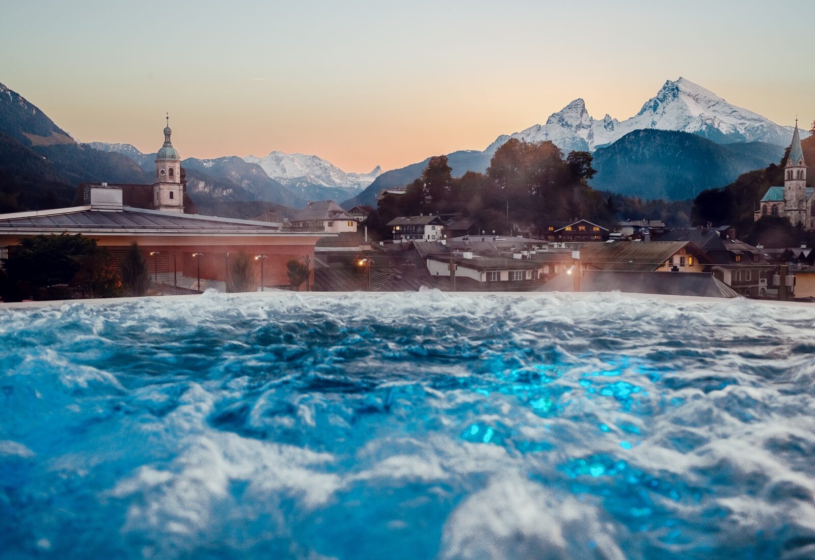 Blick auf eine Stadt mit Gebäuden und Kirchen vor einer Bergkulisse mit schneebedeckten Gipfeln im Sonnenuntergang, aufgenommen aus einem Whirlpool mit bläulichem Wasser im Vordergrund.