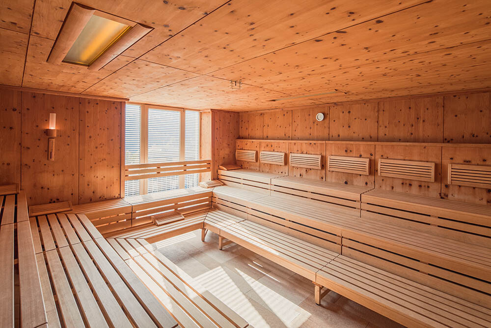 Ein Spa- oder Saunaraum mit Holzwänden, Decke und lange Bänke entlang der Wände, große Fenster, die natürliches Licht hereinlassen, und eine kleine Teppichmatte auf dem Boden.