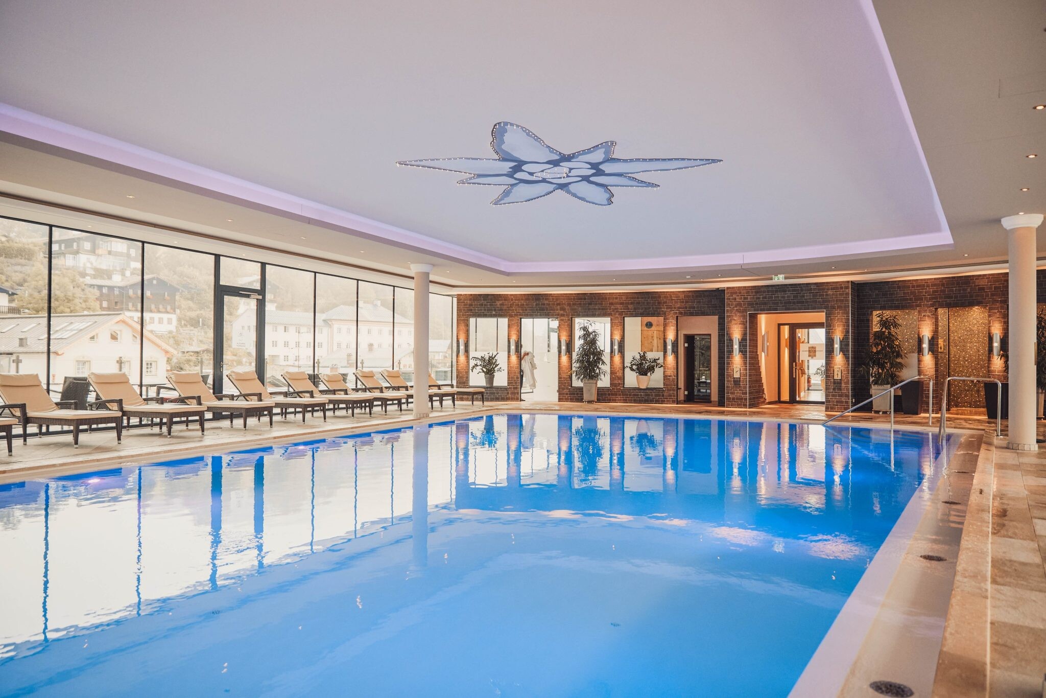 Innenaufnahme eines schwimmbads mit blauer Wasseroberfläche, umgeben von Liegestühlen und großen Fenstern, die den Blick auf eine Stadt mit Hügeln im Hintergrund bieten, und einer Deckenlila-Umrahmung mit dekorativem Licht.