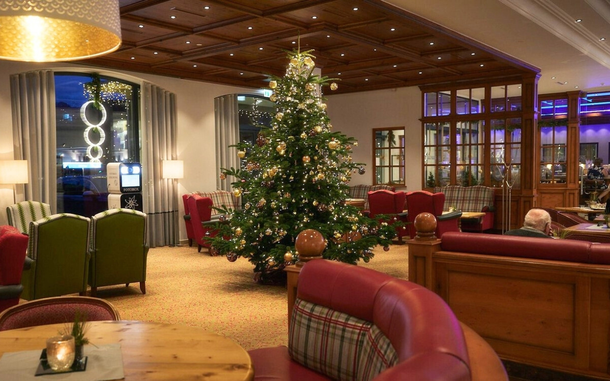 A festively decorated Christmas tree in the lobby of Hotel EDELWEISS Berchtesgaden, surrounded by cozy armchairs and tables, adorned with decorations and warm lighting.