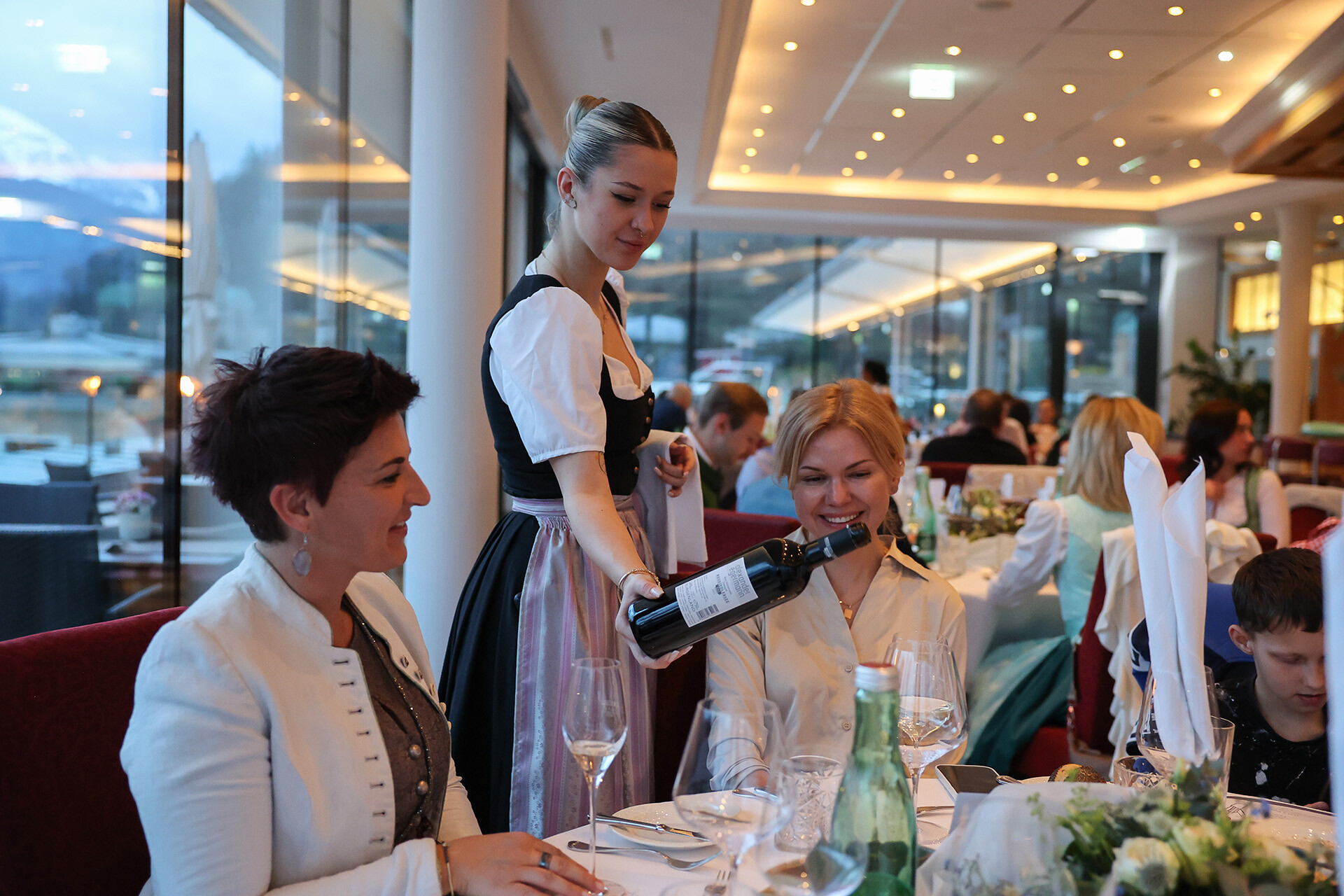 Auszubildende serviert Wein an Gäste und Hotelchefin während des Abendessens im Restaurant Panorama