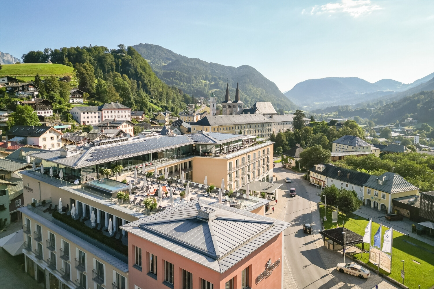 Modern hotel complex with a rooftop terrace and sun umbrellas, set within a picturesque townscape and surrounded by mountains in the background.