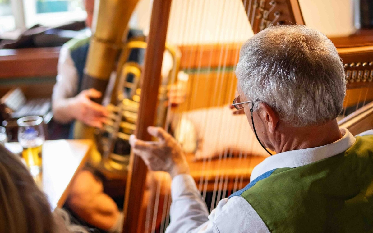 Ein älterer Mann mit grauem Haar und Brille spielt eine große goldene Harmonika, während andere Personen in einem gemütlichen Raum sitzen, mit einem Glas Bier auf dem Tisch.