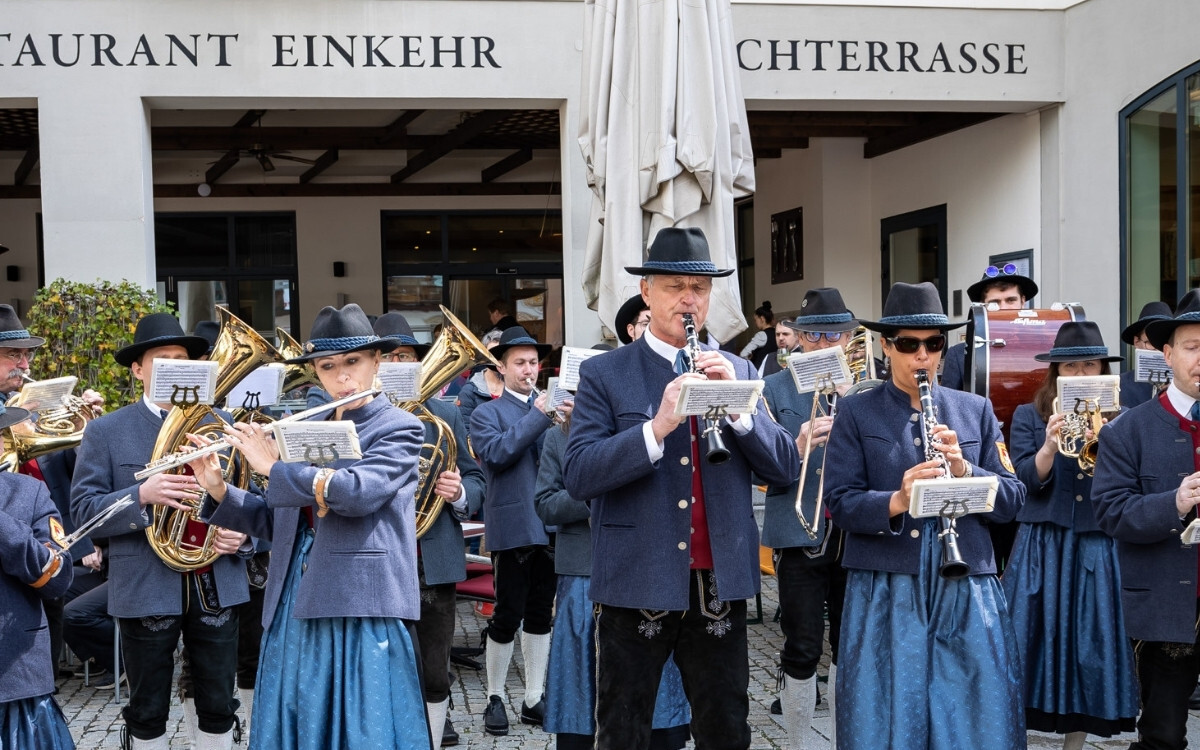 Eine Gruppe von Musikern in traditionellen Trachten, die auf einem Platz vor einem Restaurant namens 'Einkehr' mit offener Terrasse spielen.