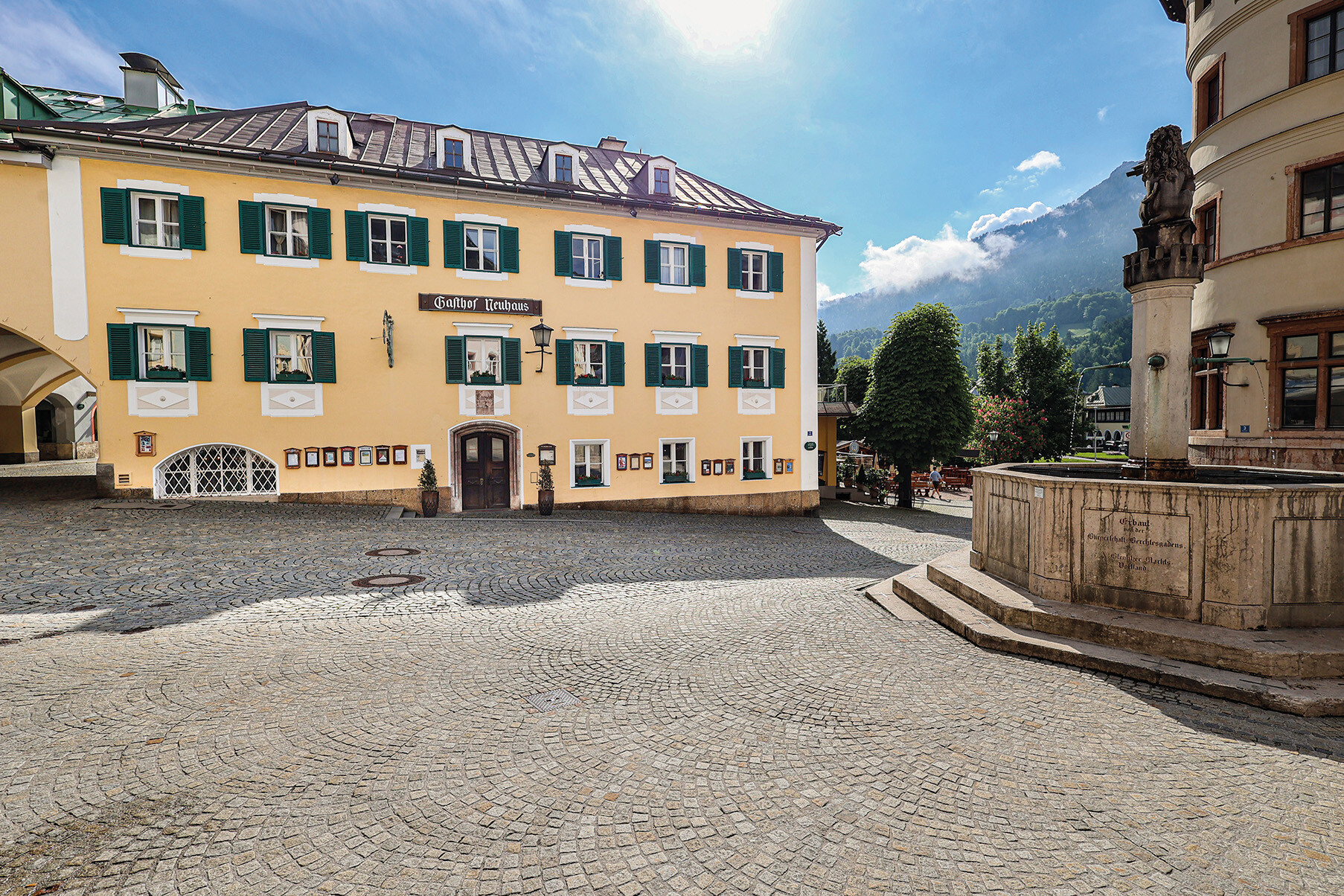 Das Bild zeigt ein ruhiges, gepflastertes Stadtplätzchen mit einem gelben Gebäude mit grünen Fensterläden und einem Schild "Gasthof Neuehaus", umgeben von Bäumen und Bergen.