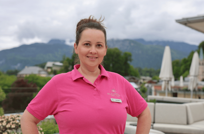 Frau in pinkem Polo-Shirt mit Namensschild steht draußen mit Bergen und bewölktem Himmel im Hintergrund.