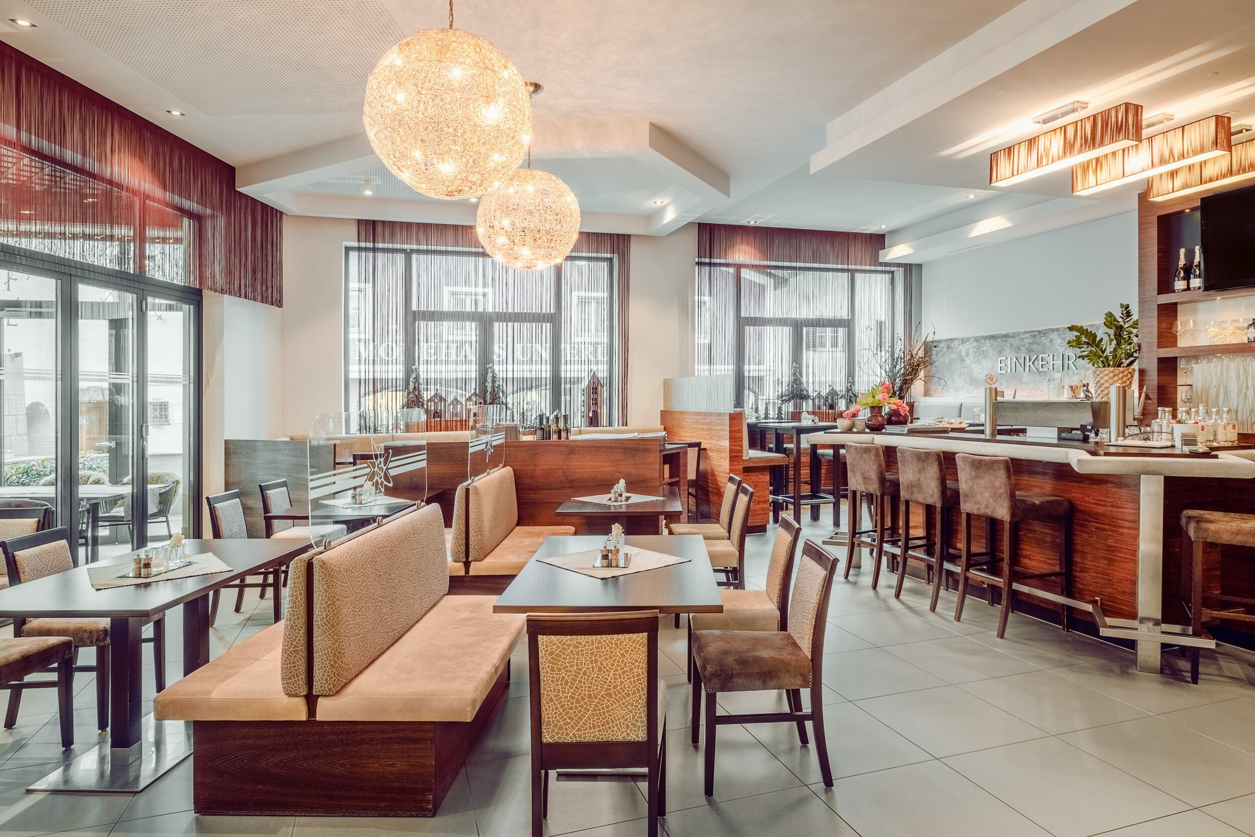 Innenaufnahme eines modern eingerichteten Restaurants mit Sitzgelegenheiten aus Holz und Stoff, großen Fenstern, dekorativen Lampen und einem Barbereich auf der rechten Seite.