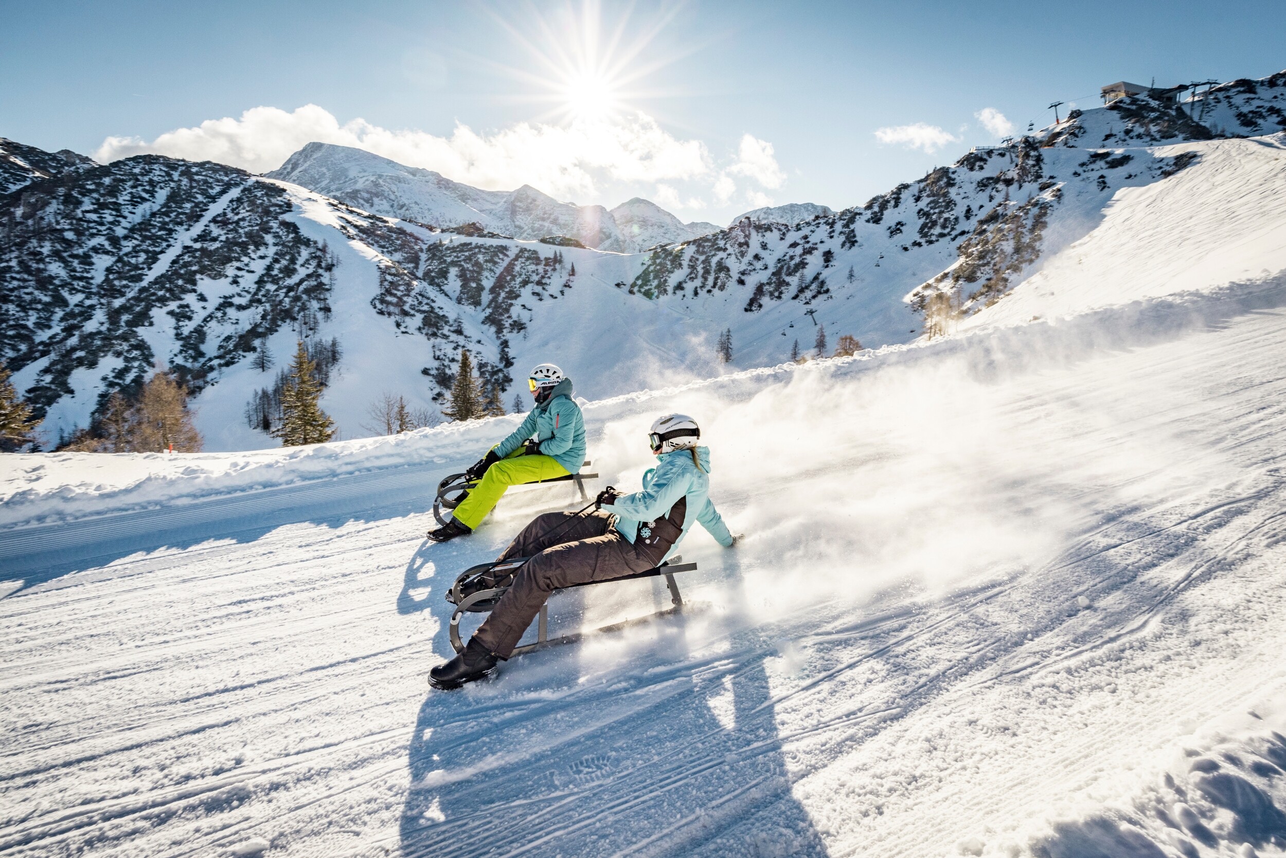 Two people enjoying a fast downhill ride on snow bikes on a sunny alpine slope surrounded by snowy mountains.