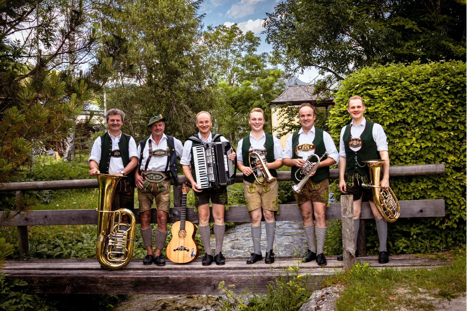 Sieben Männer in traditionellen Trachten stehen auf einer Holzbrücke im Grünen, jeder hält ein Musikinstrument, darunter Saxophon, Akkordeon, Trompete, Tuba und Horn. Auzinger Tanzlmusi