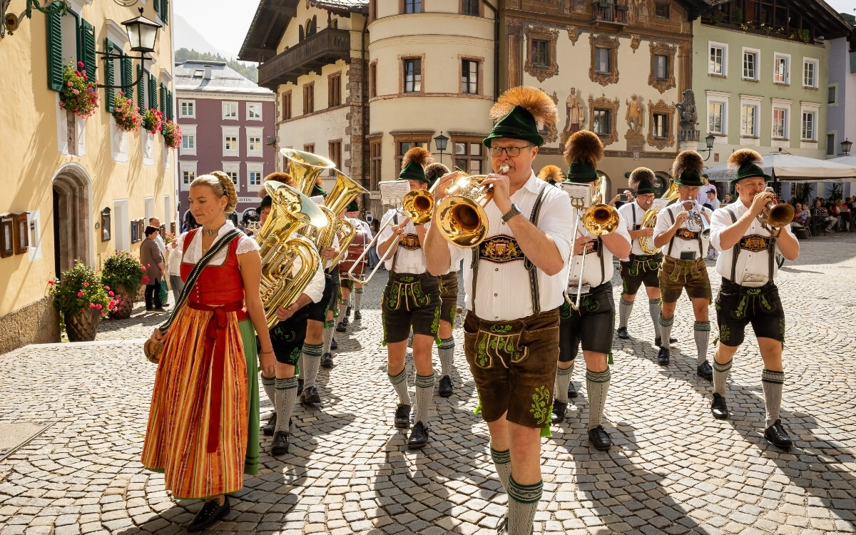 Eine Gruppe von Musikern in traditioneller bayerischer Kleidung, die auf einem gepflasterten Platz in Berchtesgaden vor dem Gasthof NEUHAUS mit bunten Fassaden und Blumenarrangements auf den Fenstern marschiert, während sie Blasinstrumente spielt.