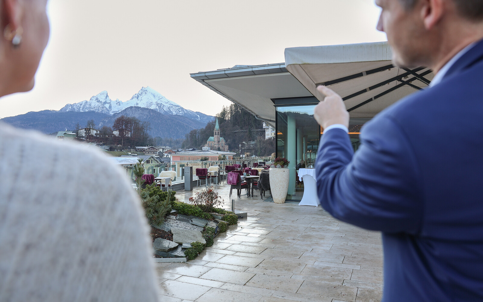 Hoteldirektor Christop Schimpl im Gespräch auf einer Terrasse mit Blick auf Berchtesgaden und den Watzmann im Hintergrund, eine Person zeigt mit dem Finger auf das Gebäude.